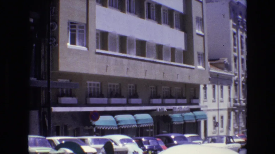1972 Portugal street frame with parked cars, a light-colored car crossing the lower foreground, teal awnings, and a multi-story beige building behind them.