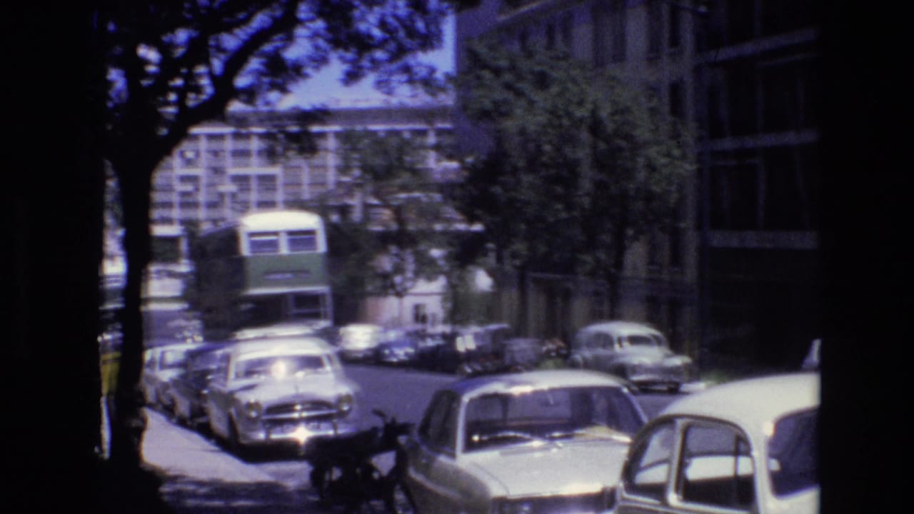 Portugal 1972 8mm frame showing a green double-decker bus moving toward the camera on a tree-lined urban street with parked cars and modern buildings.