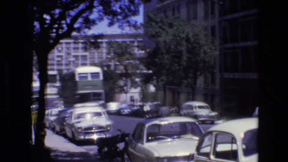 Portugal 1972 8mm frame showing a green double-decker bus moving toward the camera on a tree-lined urban street with parked cars and modern buildings.
