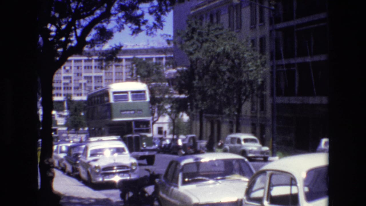 Portugal 1972 8mm frame with a green double-decker bus traveling closer through a city street bordered by parked sedans, trees, and tall modern buildings.