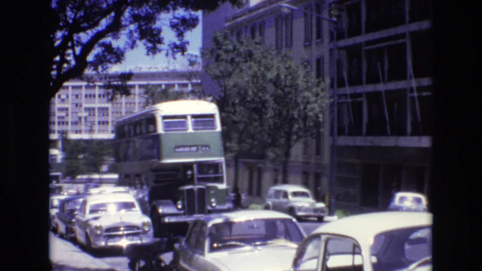 Portugal 1972 8mm frame showing the double-decker bus nearer to camera, with a row of parked cars, a pale car in the foreground, and modern buildings along the street.