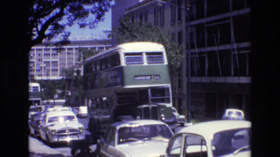 Portugal 1972 8mm frame with the green double-decker bus filling more of the view as it moves along a tree-lined street with parked cars and office-like buildings.