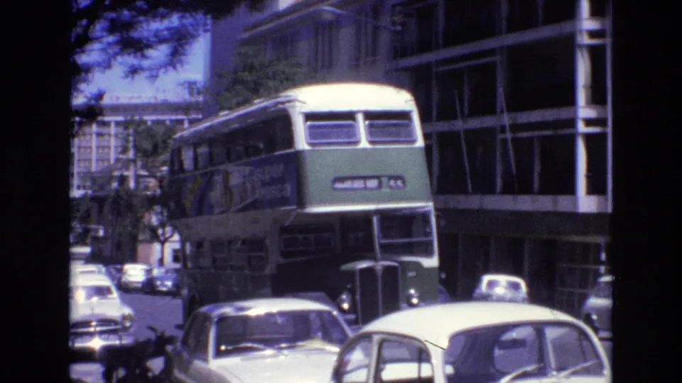 A green and cream double-decker bus approaches on a Portugal city street in footage identified as 1972, with compact cars, buildings, and trees around it.