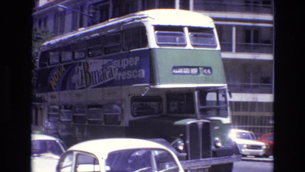 The double-decker bus moves closer through traffic, showing a large Nova Binaca super fresca advertisement along its side in the 1972 Portugal street scene.