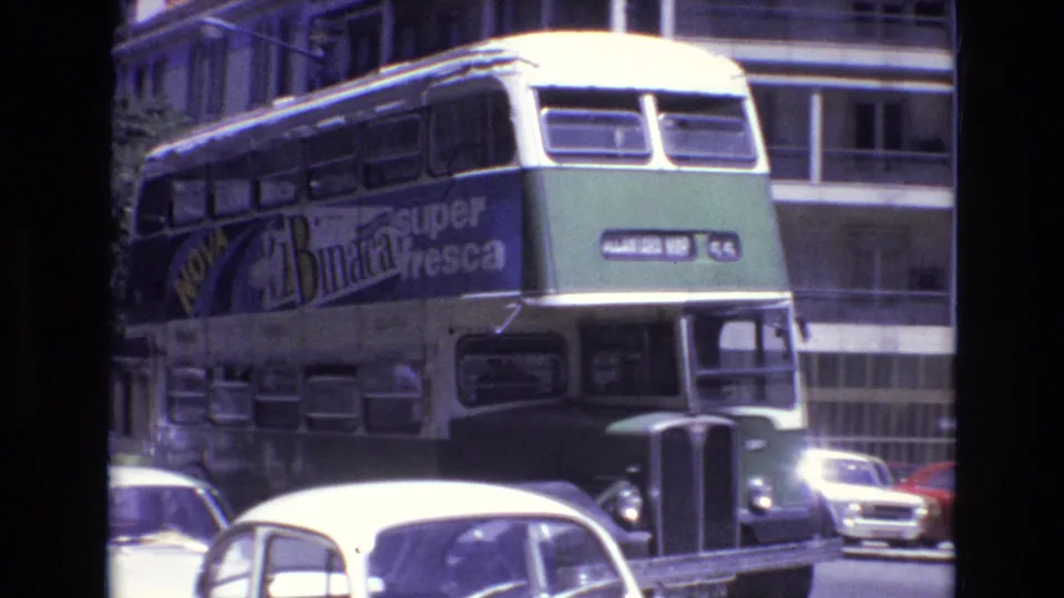 The double-decker bus moves closer through traffic, showing a large Nova Binaca super fresca advertisement along its side in the 1972 Portugal street scene.