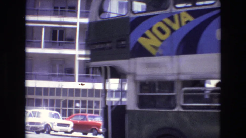 1972 Portugal street view with a green and cream double-decker bus carrying a NOVA advertisement beside apartment buildings and parked compact cars.