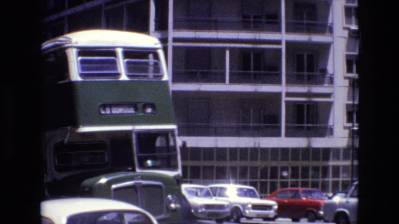 Front view of a green and cream double-decker bus in Portugal in 1972, framed against apartment balconies and parked compact cars.
