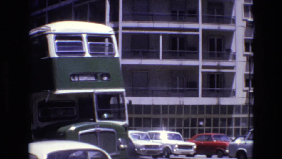 Front view of a green and cream double-decker bus in Portugal in 1972, framed against apartment balconies and parked compact cars.
