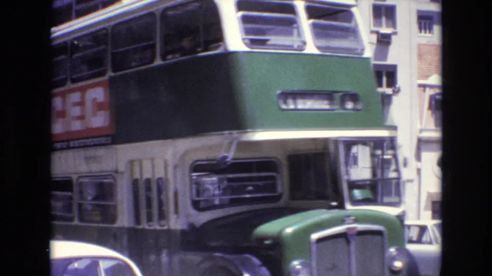 Close 1972 Portugal street frame of a green and cream double-decker bus passing near the camera, with advertising panels and parked cars partly visible.