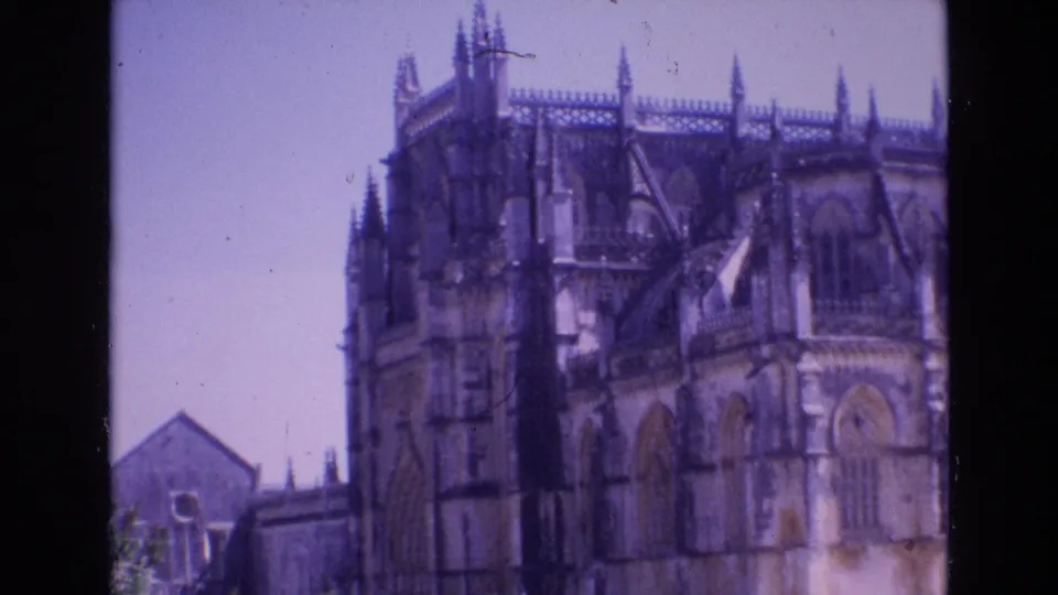 1972 Portugal 8mm frame showing a historic stone church facade with Gothic-style arches, roof pinnacles, and soft purple-blue film color.