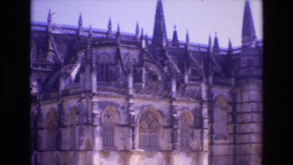 1972 Portugal 8mm frame showing a wider portion of the historic church exterior, including arched windows, roof pinnacles, a tall spire, and adjoining stone structure.