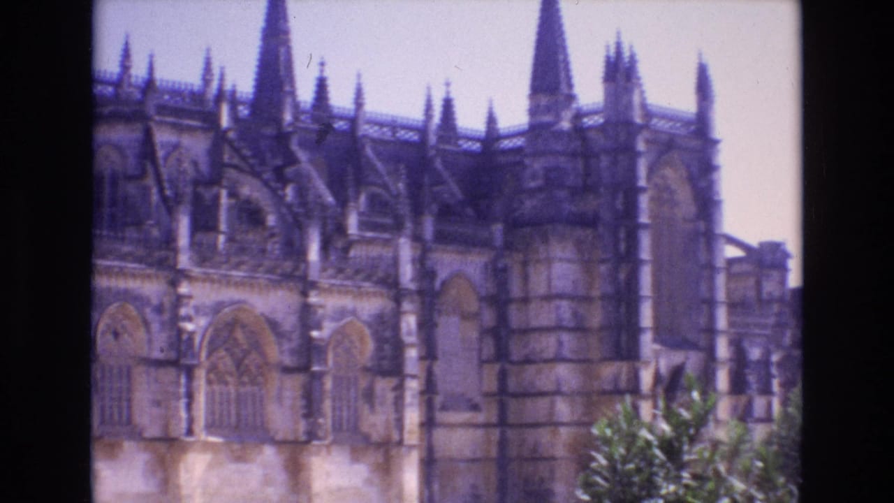 1972 Portugal 8mm frame of a Gothic cathedral exterior, showing arched windows, vertical stonework, spires, and greenery in the lower right.