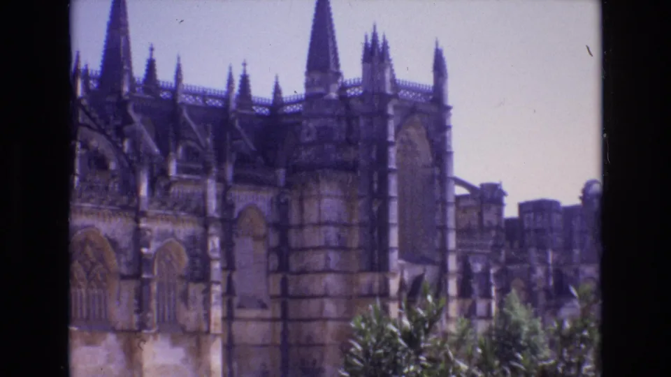 1972 Portugal 8mm frame showing a closer visitor-shot view of the cathedral's arched stone facade, tower-like corner, spires, and foreground greenery.