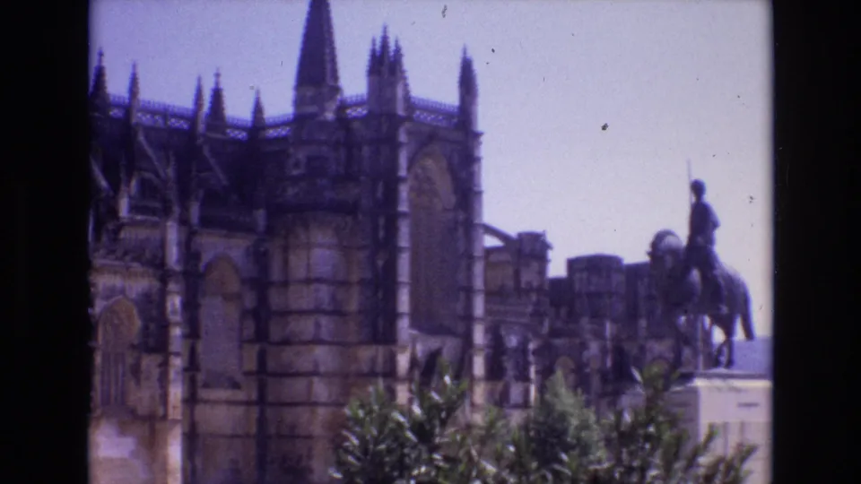 1972 Portugal 8mm frame showing the cathedral exterior with Gothic stonework, foreground shrubs, background buildings, and a mounted statue appearing at the right edge.