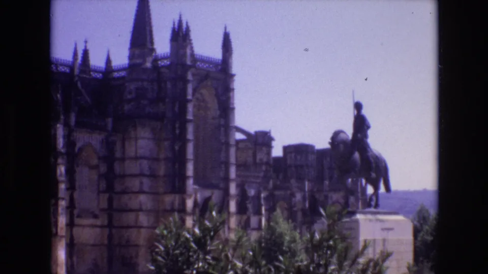1972 Portugal 8mm frame showing a rider-on-horseback statue in front of a Gothic-style stone building, with foliage in the foreground and pale sky behind.
