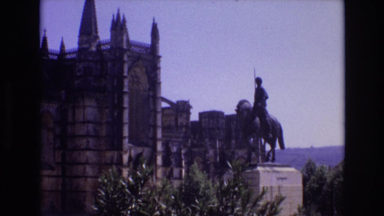 1972 Portugal home-movie frame with the equestrian monument centered to the right of a dark Gothic-style facade and towers, framed by shrubs below.