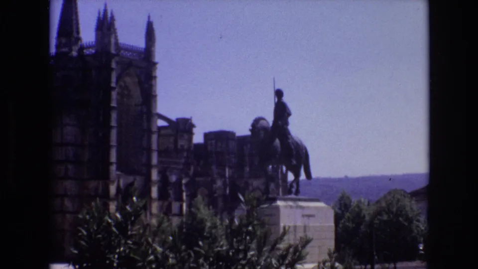 1972 Portugal travel frame with a rider statue silhouetted against the sky near Gothic-style towers, foreground shrubs, and a distant horizon beyond the architecture.