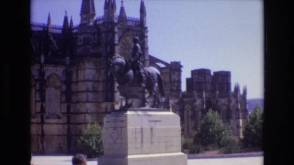 1972 Portugal home-movie frame showing the equestrian statue beside Gothic cathedral stonework, with shrubs, plaza pavement, and a cropped visitor near the foreground edge.