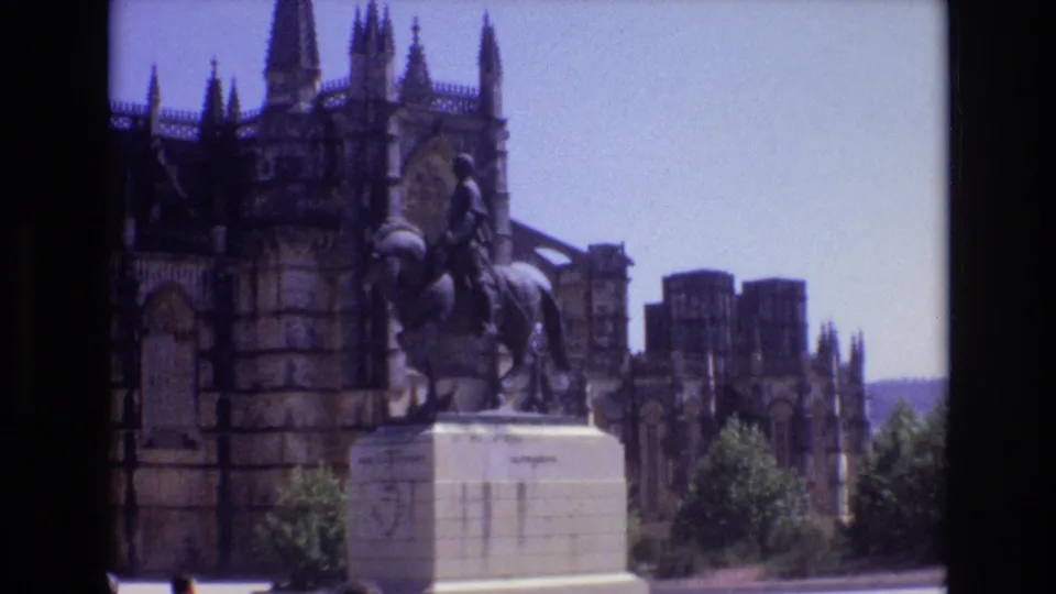 1972 Portugal 8mm frame showing the equestrian monument set before a dark Gothic cathedral facade, with trees, bright pavement, and distant hills at the side.