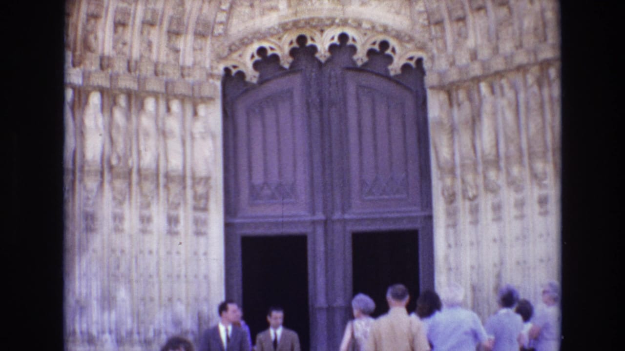Visitors in 1970s attire stand at the large wooden doors of a carved cathedral entrance in Portugal.