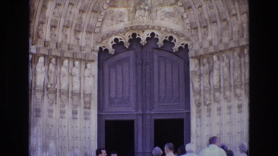 Closer 8mm frame of the ornate cathedral portal and tall wooden doors as visitors gather and move near the entrance.