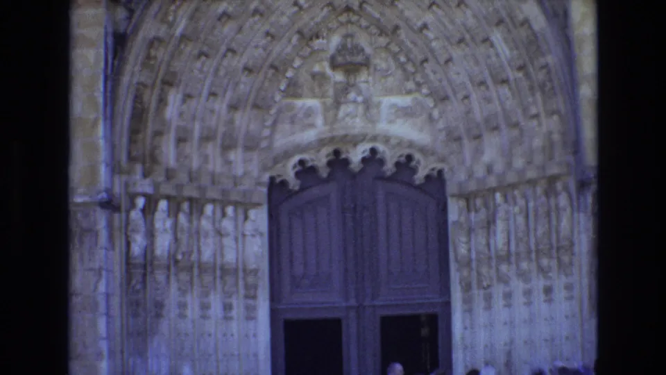 1972 Portugal 8mm frame showing an ornate Gothic church portal with carved stone arches, statues, large dark wooden doors, and partially visible visitors near the entrance.