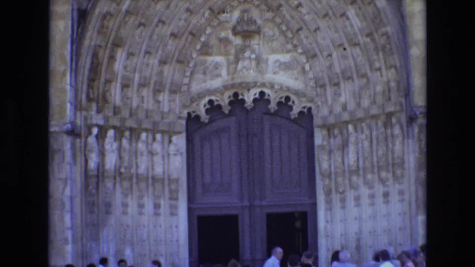 1972 Portugal 8mm frame of a Gothic cathedral entrance with ornate stone carving, dark double doors, and visitors standing or moving along the base of the facade.