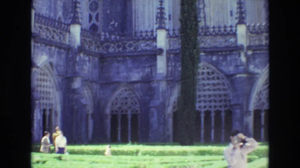 1972 Portugal 8mm frame showing a sunlit cathedral courtyard with green lawn, Gothic stone windows and arches, a tall narrow tree, and visitors walking or taking photographs.