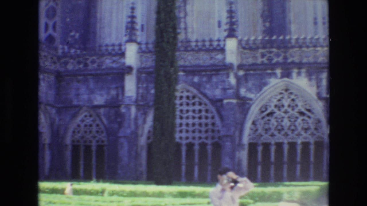 1972 Portugal 8mm frame showing a Gothic-style building facade with pointed arched windows, clipped hedges, a tall tree trunk, and a tourist raising a camera near the lower edge.