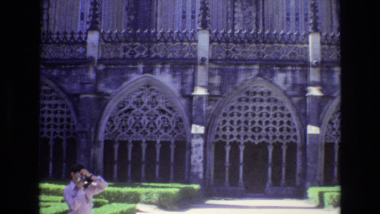 1972 Portugal 8mm frame with an adult tourist holding a camera in front of ornate Gothic-style arched windows, stone columns, clipped hedges, and a bright walkway.