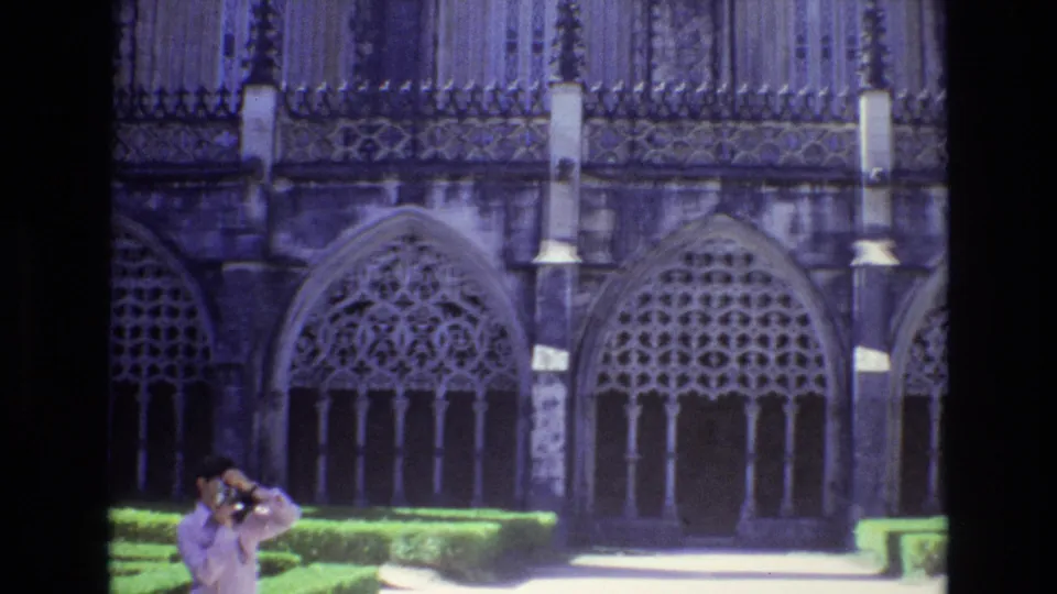 1972 Portugal 8mm frame with an adult tourist holding a camera in front of ornate Gothic-style arched windows, stone columns, clipped hedges, and a bright walkway.