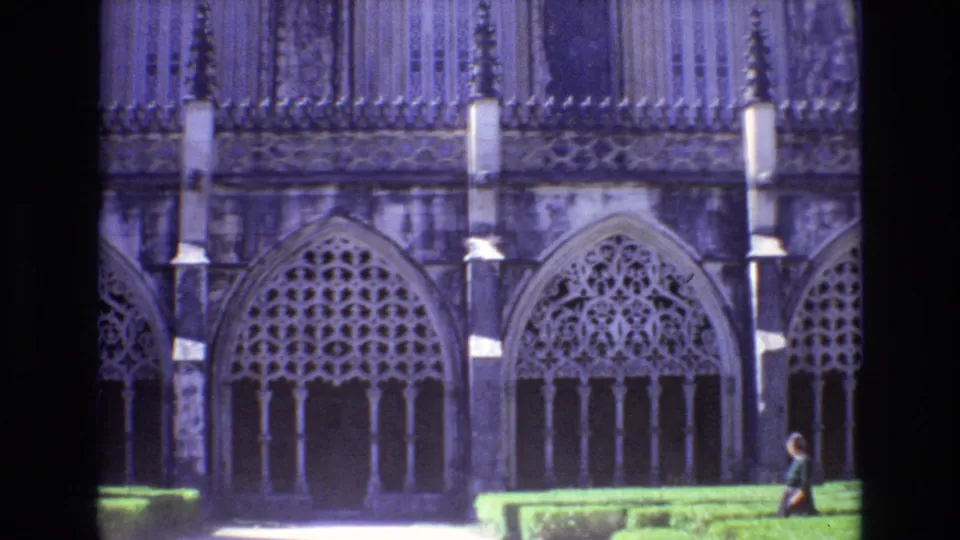 1972 Portugal 8mm frame showing a Gothic-style stone facade with repeated pointed arches, clipped hedges, a bright path, and a small visitor walking at frame right.