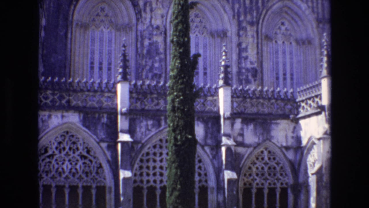 1972 Portugal 8mm close-up of ornate Gothic-style stone arches, tracery windows, and a tall cypress tree in front of a religious building.