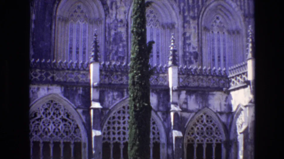 1972 Portugal 8mm close-up of ornate Gothic-style stone arches, tracery windows, and a tall cypress tree in front of a religious building.