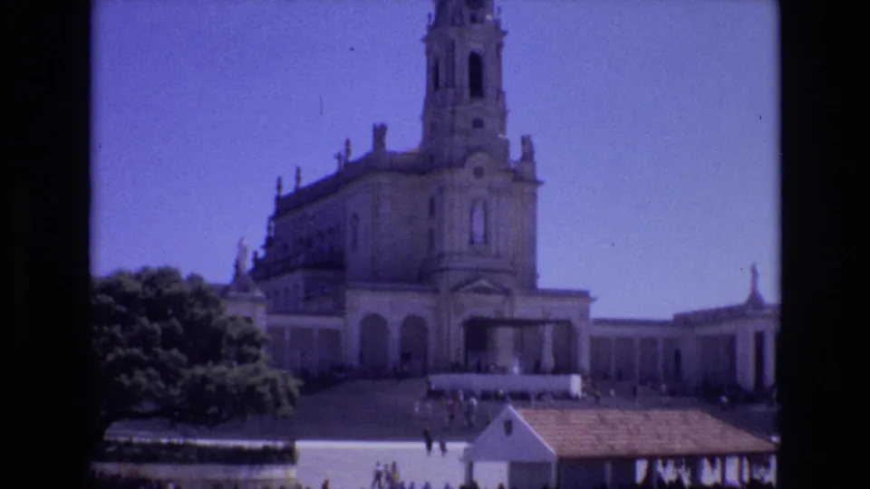 Wide 8mm view in Portugal of a pale stone religious building with a tower, flanking structures, trees, and distant visitors under a clear blue sky.