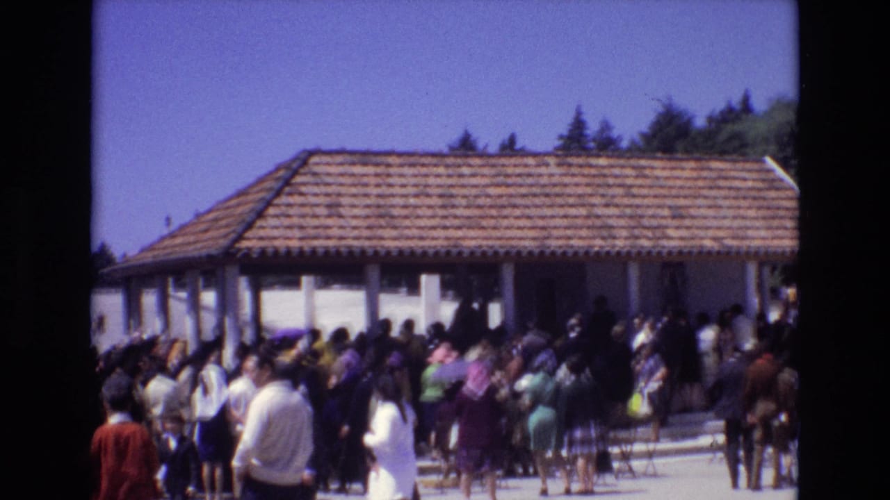 People in 1970s attire gather beside a white pavilion with a tiled roof in Portugal, filmed from a handheld home-movie viewpoint.
