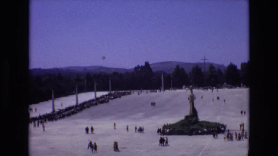 Small groups cross a broad sunlit plaza in Portugal as a line of visitors extends toward a central statue or monument with wooded hills in the background.