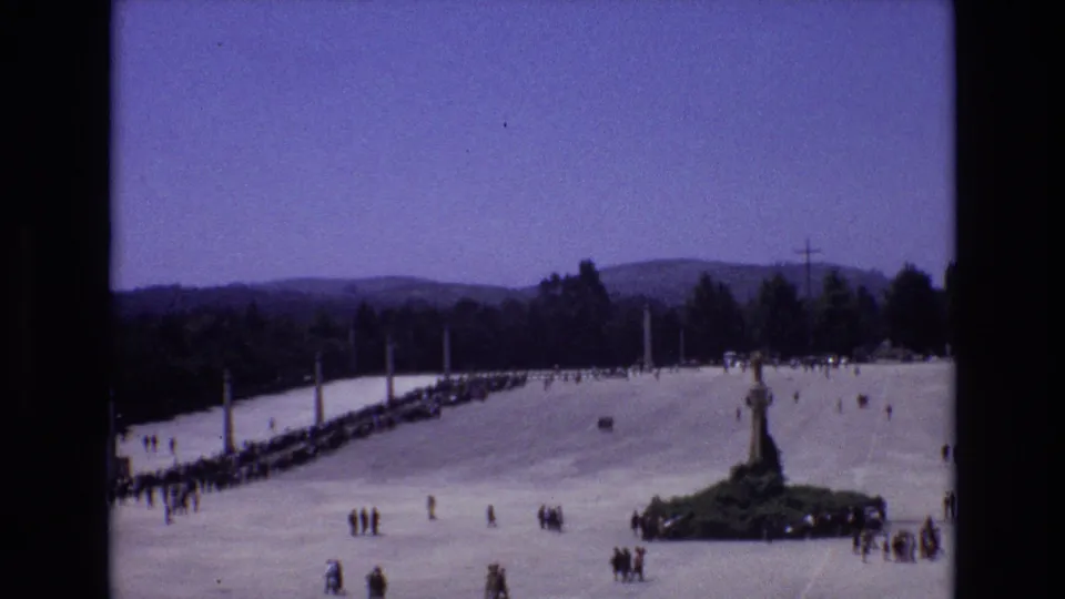 A handheld 8mm view looks across the plaza toward a central monument, with scattered visitors, trees, hills, and a bright blue sky visible.