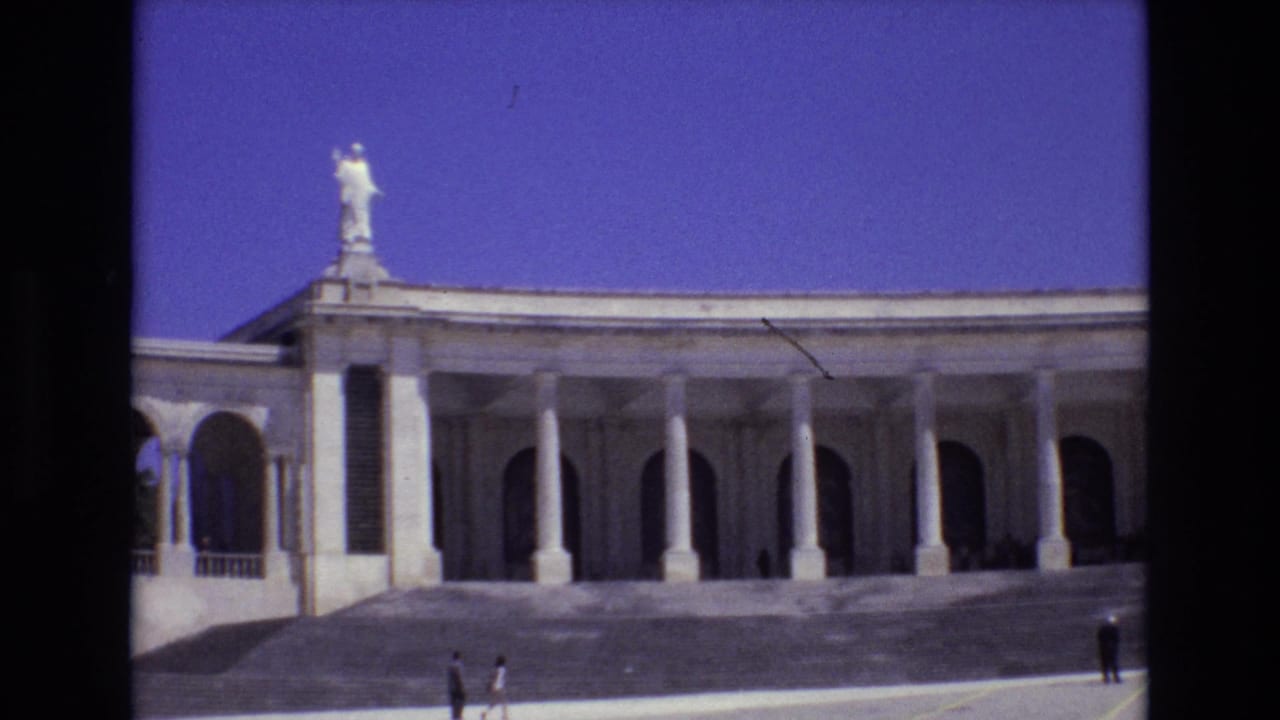 1972 Portugal 8mm frame showing a long white colonnaded building with arched openings, a rooftop statue, blue sky, and distant visitors in the plaza.