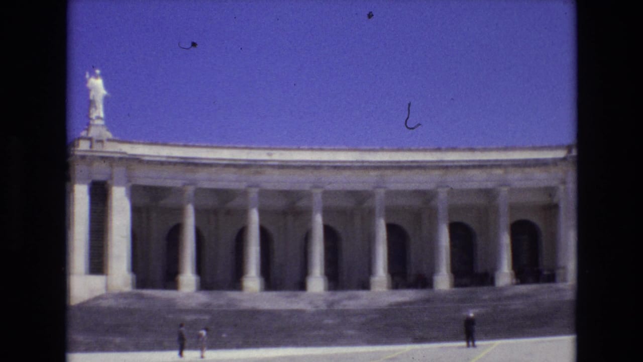 1972 Portugal travel footage frame with a monumental colonnade, repeated columns, arched bays, a white rooftop statue, and small sightseeing figures below.