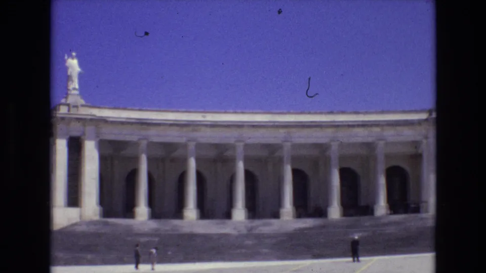 1972 Portugal travel footage frame with a monumental colonnade, repeated columns, arched bays, a white rooftop statue, and small sightseeing figures below.
