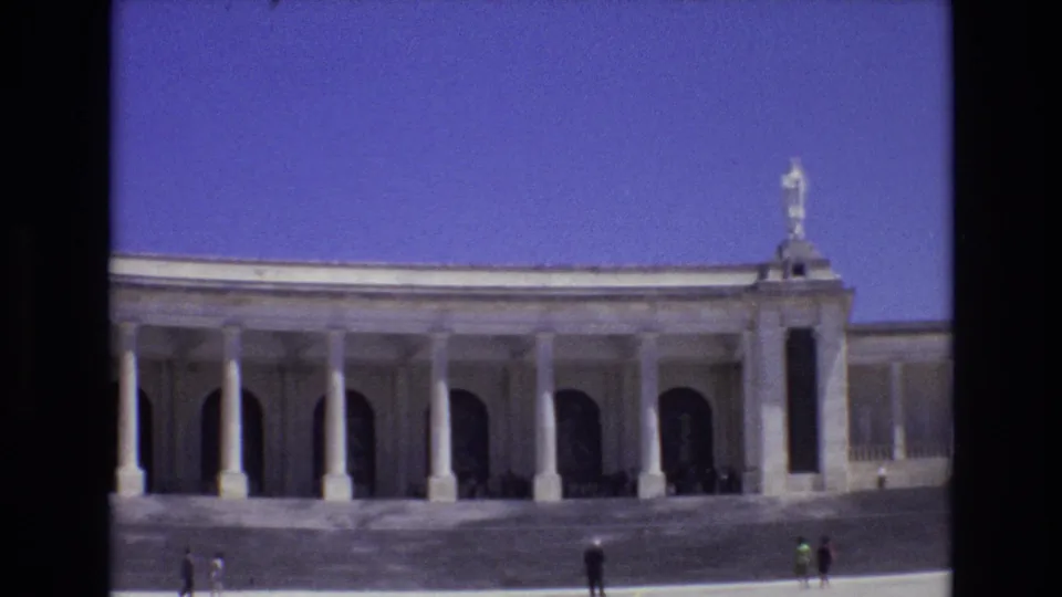 1972 Portugal home-movie frame showing a neoclassical-looking colonnaded facade, dark archways, a rooftop statue, and distant visitors moving across the foreground.