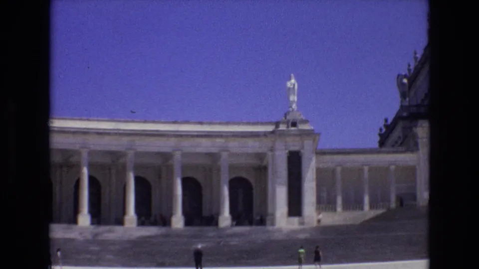 1972 Portugal 8mm frame from a slightly shifted angle, showing the white colonnade, rooftop statue, adjoining building edge, open plaza, and distant visitors.