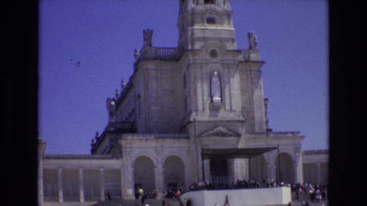1972 Portugal 8mm frame showing a pale stone cathedral facade with a tall bell tower, arched entrance area, distant visitors, and clear blue sky.