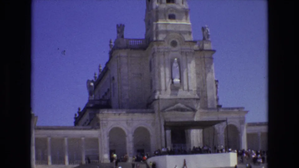 1972 Portugal 8mm frame showing a pale stone cathedral facade with a tall bell tower, arched entrance area, distant visitors, and clear blue sky.