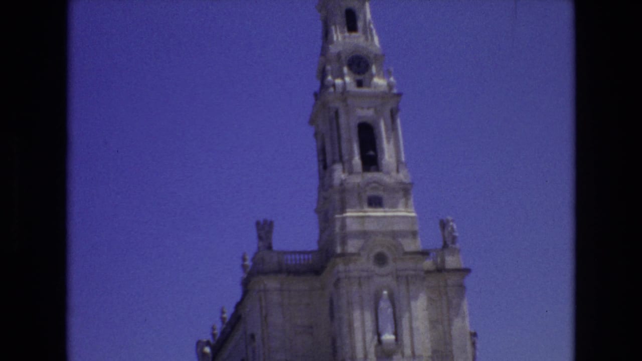 1972 Portugal 8mm frame showing the upper section of a pale stone cathedral tower with arched openings and clock detail against a clear blue sky.