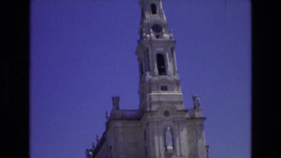 1972 Portugal 8mm frame showing the upper section of a pale stone cathedral tower with arched openings and clock detail against a clear blue sky.