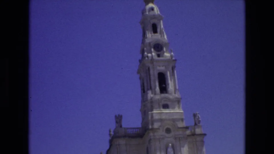 1972 Portugal home-movie frame with the cathedral tower positioned to the right, showing the clock face, arched openings, and sculpted stonework under blue sky.