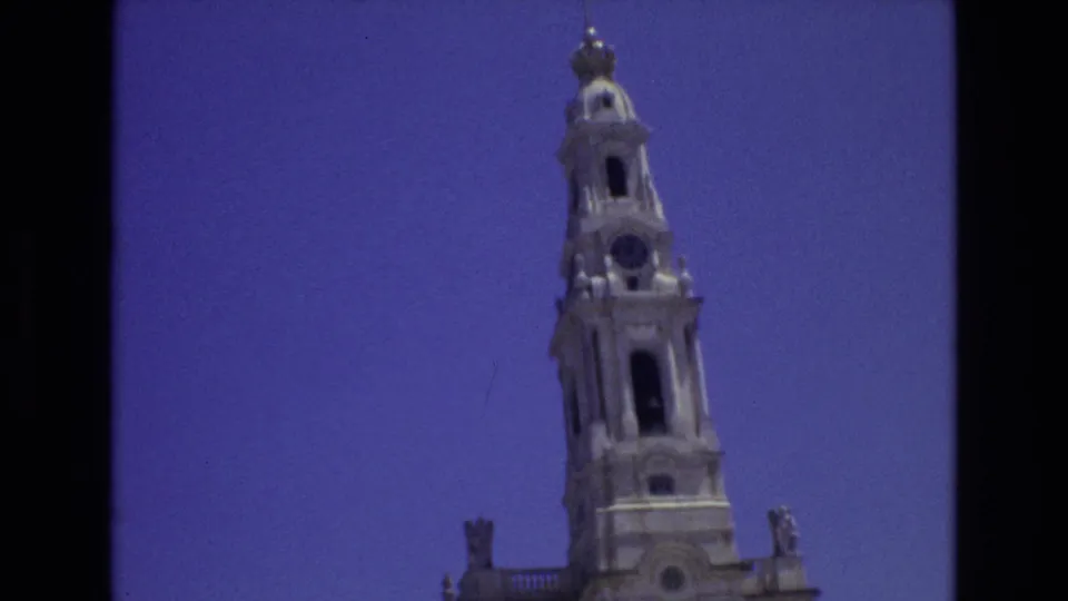 1972 Portugal 8mm frame showing a closer upward view of the cathedral tower's spire, clock face, arched bell opening, and stone ornamentation.