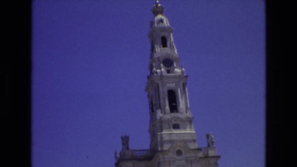 1972 Portugal frame of the cathedral tower centered in the home-movie shot, with a domed spire, clock face, arched openings, and blue sky visible.